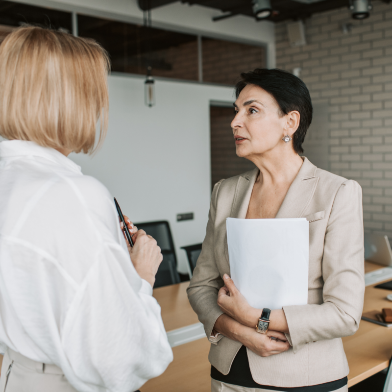 Dos mujeres ejecutivas conversando en una sala corporativa moderna, representando liderazgo intergeneracional, experiencia y relevancia profesional dentro del nuevo mundo laboral.